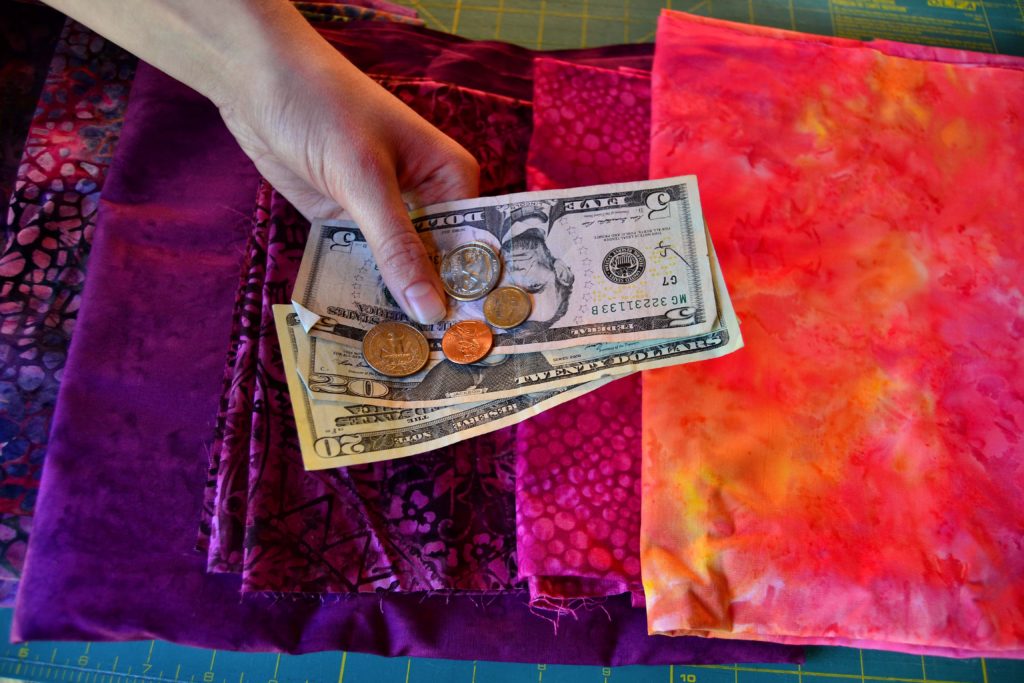 Handing holding cash and coin money over purple and pink quilting fabrics sewing within your means illustration